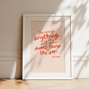 Puede incluir: Una impresión enmarcada en blanco con un fondo rosa y texto rojo que dice "The cure for anything is salt water: sweat, tears or the sea - ISAK DINESEN".