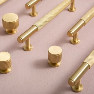 Könnte beinhalten: Ein Satz von fünf goldfarbenen Schrankknöpfen und -griffen mit geriffeltem Design. Die Knöpfe sind rund und die Griffe sind T-förmig.