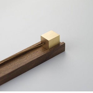 Puede incluir: Un porta incienso de madera con un cuadrado de latón en la parte superior. El soporte está hecho de madera oscura y tiene un diseño simple.