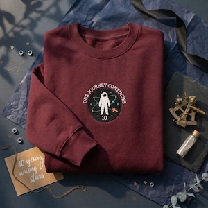 May include: A maroon crewneck sweatshirt featuring a circular embroidered patch on the chest. The patch depicts a white astronaut, colorful planets, and the text "OUR JOURNEY CONTINUES" with the number "10" centered at the bottom.