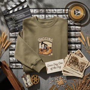 Puede incluir: Una sudadera de cuello redondo color verde oliva con un bordado que muestra una bota de trabajo y una torre de perforación petrolífera con el texto "DIGGING" encima. La sudadera se muestra junto a una claqueta, rollos de película y fotos antiguas.
