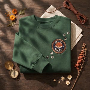 May include: A forest green crewneck sweatshirt featuring a circular patch on the chest with a fox illustration and the text AMAZE. The design includes embroidered paw prints leading toward the patch, which is set against a dark fabric background.