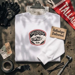 May include: A white long-sleeve sweatshirt featuring a central embroidered patch with a race car, smoke, and the text "TALLADEGA MADE ME" and "77". The design includes checkered flags and a red border, centered on the chest of the garment.