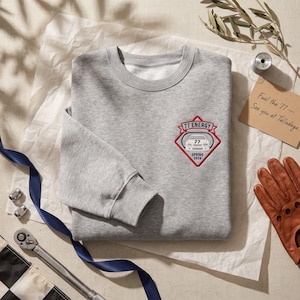 May include: A heather gray crewneck sweatshirt featuring an embroidered patch on the left chest. The patch is a red and white diamond shape with a race car graphic, the text 77 ENERGY, and the text SPRING 2026 centered within the design.