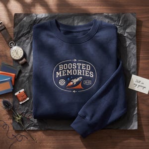 May include: A navy blue crewneck sweatshirt featuring an embroidered oval design on the chest. The graphic includes the text "BOOSTED MEMORIES" above a rocket ship, a checkered flag, and the number "10.01" inside a rectangular border.