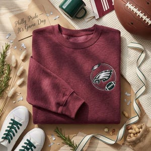 Könnte beinhalten: Ein kastanienbraunes Sweatshirt mit Rundhalsausschnitt und einem gestickten Philadelphia Eagles-Logo auf der linken Brust, das einen Adlerkopf und einen Football in einem kreisförmigen Rahmen zeigt. Es hat lange Ärmel und gerippte Bündchen.