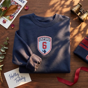 May include: A navy blue crewneck sweatshirt featuring a central embroidered shield patch. The patch displays the text OXFORD in white, the number 6 in red with a white outline, and a small silhouette of a football player in navy blue.