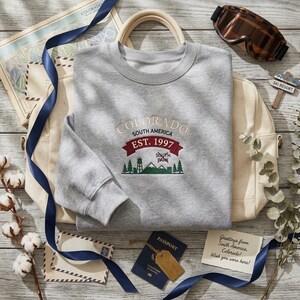 Puede incluir: Una sudadera gris jaspeado de cuello redondo con texto bordado que dice Colorado South America Est. 1997, junto a un gráfico de montañas, pinos y una torre de agua. El diseño incluye una cinta roja y detalles en color verde.