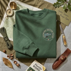 May include: A forest green crewneck sweatshirt featuring a circular embroidered patch on the left chest. The patch displays a stylized alligator head graphic with the text "LEONIDA GATOR CLUB" and a small star icon in a circular border.