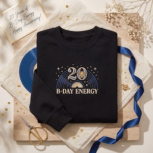 May include: A black long-sleeve sweatshirt featuring an embroidered design with the number 20, a bee, and the text B-DAY ENERGY. The graphic includes blue concentric circles resembling a vinyl record, gold stars, and confetti accents.