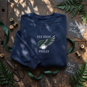 Pode incluir: Uma camisola azul-marinho de gola redonda com um design bordado de uma bola de futebol americano branca com asas verde-escuras. O texto "FLY HIGH" está posicionado acima da bola e "PHILLY" está bordado abaixo numa fonte branca.