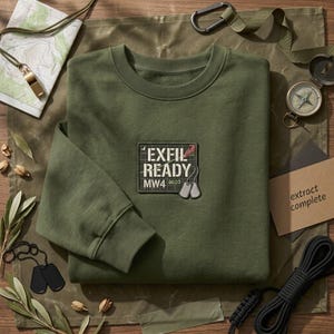 Könnte beinhalten: Ein olivgrünes Sweatshirt mit Rundhalsausschnitt und einem zentralen gestickten Aufnäher mit dem Text EXFIL READY MW4 00:23, ergänzt durch eine Grafik von zwei silbernen Erkennungsmarken. Das Sweatshirt hat lange Ärmel mit Rippbündchen.