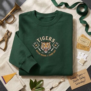 May include: A dark green crewneck sweatshirt featuring an embroidered graphic of a tiger face centred between two football goalposts. The text reads TIGERS, BATON ROUGE, and GAME DAY. The design includes small football icons and stadium lights.
