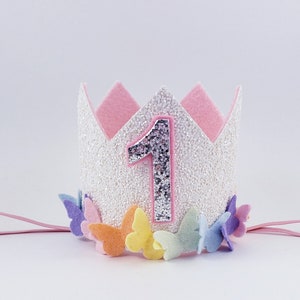 May include: A white glitter crown with a pink felt top and a silver felt number one. The crown is decorated with colorful felt butterflies and has a pink elastic band.