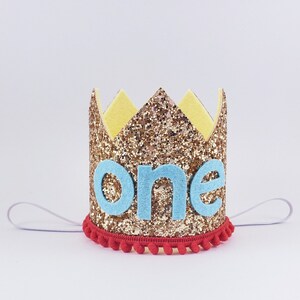 May include: Gold glitter crown with a blue felt "one" on the front. The crown has a red pom-pom trim and a white elastic band.