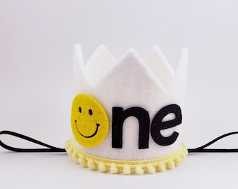 May include: White felt crown with a black felt "one" and a yellow smiley face. The crown has a black elastic band and yellow pom-poms around the base.