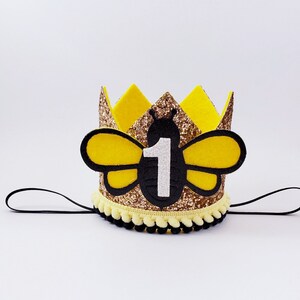 May include: A gold glitter bee-themed crown with black felt wings and a white felt number "1" on the front. The crown has a black elastic band for wearing.