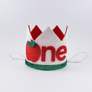 May include: White felt crown with red felt apple and green felt leaf, and the word "one" in red felt letters. The crown has a white elastic band.