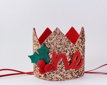 Könnte beinhalten: Goldene Glitzerkrone mit roter Filzbasis und einem roten Filz "eins" auf der Vorderseite. Die Krone hat ein grünes Filz-Stechpalmenblatt mit roten Filzbeeren an der Seite. Die Krone hat eine rote Filzschlaufe.