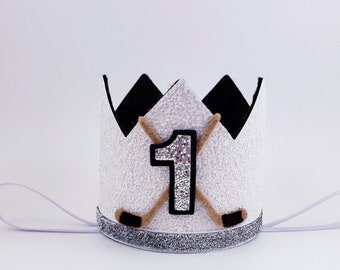 May include: White felt crown with a silver glitter trim, featuring a black felt hockey stick and a brown felt hockey stick crossed behind a large silver glitter number one.