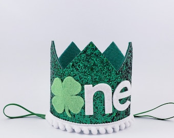 May include: Green glitter crown with a white felt shamrock and the word "one" in white felt. The crown has a white pom-pom trim and a green felt tie.