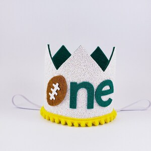 May include: White felt crown with a green felt trim and a brown felt football. The crown has yellow pom-poms around the bottom and says "#one" in green felt letters.