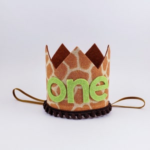 May include: A brown felt crown with a giraffe print and a green felt "one" on the front. The crown has a brown felt band with brown pom-poms around the edge.