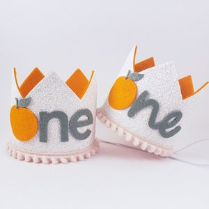May include: Two white felt crowns with orange felt fruit and green felt leaves. The crowns have a pink pom-pom trim and say "one" in gray felt letters.