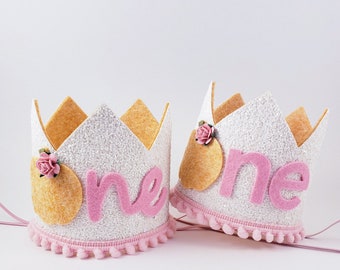 May include: Two white glitter crowns with pink felt trim and pink felt "one" lettering. Each crown has a gold felt circle and a pink felt flower.