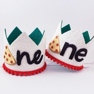 May include: Two white felt crowns with red pom-poms around the base. Each crown has a green felt pizza slice with red and yellow accents and the word "one" in black felt.