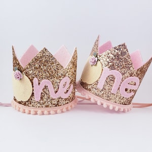 May include: Two gold glitter crowns with pink felt accents and the word "one" in pink felt letters. Each crown has a pink felt flower and a yellow felt circle.