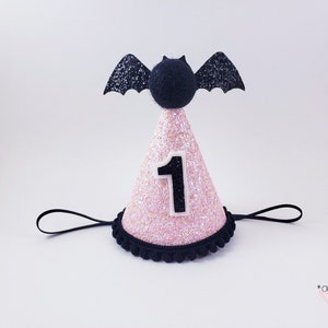 May include: Pink and black glitter party hat with a black bat on top and the number "1" on the front. The hat has a black elastic band.