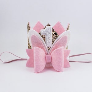May include: Pink felt crown with gold glitter, white felt bunny ears, and a white felt number one. The crown is attached to a pink elastic headband.