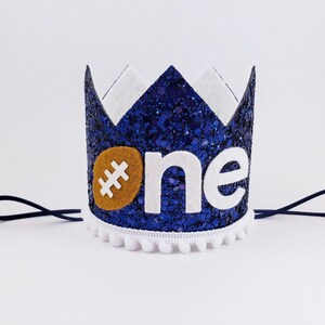 May include: Blue glitter crown with white felt trim and a white felt #one with a brown felt football. The crown has a white pom-pom trim and a black elastic band.