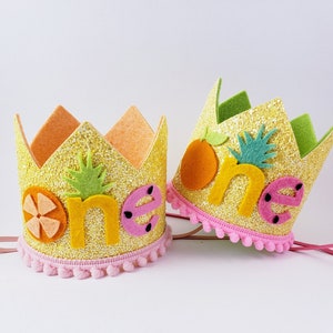 May include: Two yellow glitter crowns with pink pom pom trim. The crowns are decorated with felt fruit and the word "one" in yellow felt letters.