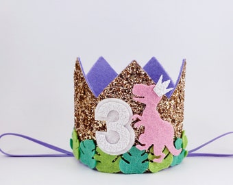 May include: A gold glitter crown with a purple felt top, a pink dinosaur with a white crown, green felt leaves, and a white felt number "3". The crown has a purple elastic band.
