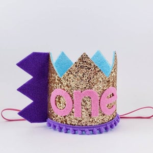 May include: Gold glitter crown with purple felt trim and pink felt "one" lettering. The crown has a purple elastic band.