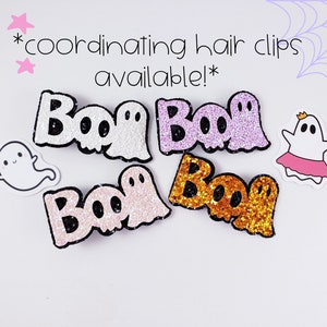 K&ouml;nnte beinhalten: Vier Glitzer-Haarklammern mit dem Wort "Boo" und einem Geisterdesign. Die Klammern sind in pink, lila, wei&szlig; und goldfarbenem Glitzer.