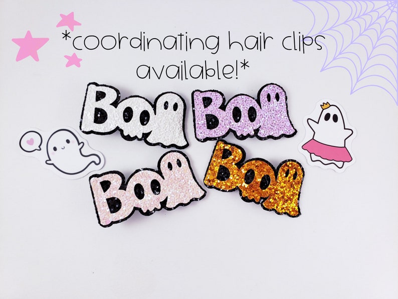 K&ouml;nnte beinhalten: Vier Glitzer-Haarklammern mit dem Wort "Boo" und einem Geisterdesign. Die Klammern sind in pink, lila, wei&szlig; und goldfarbenem Glitzer.