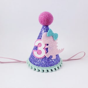 May include: Purple glitter party hat with a pink felt dinosaur, a green bow, and a white number "3". The hat has a pink felt pom-pom on top and a light pink elastic band.