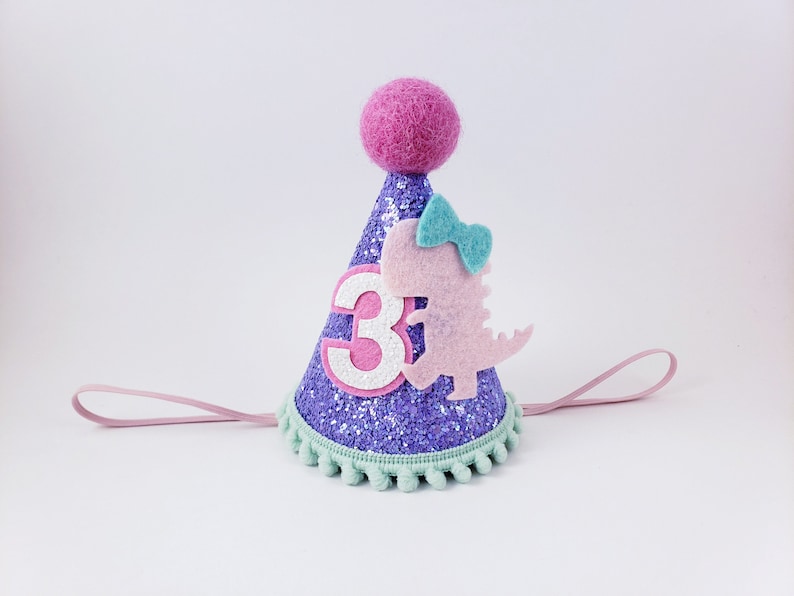 May include: Purple glitter party hat with a pink felt dinosaur, a green bow, and a white number "3". The hat has a pink felt pom-pom on top and a light pink elastic band.