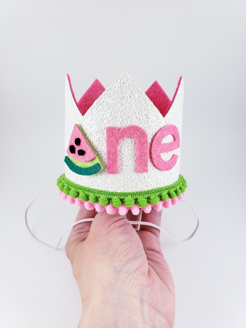 May include: White felt crown with pink felt trim and a watermelon design. The crown says "one" in pink felt letters and has a green and pink pom-pom trim.