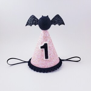 May include: Pink and black glitter party hat with a black bat on top and the number "1" on the front. The hat has a black elastic band.