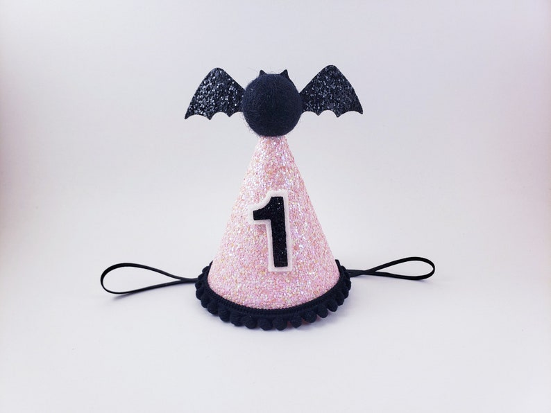 May include: Pink and black glitter party hat with a black bat on top and the number "1" on the front. The hat has a black elastic band.