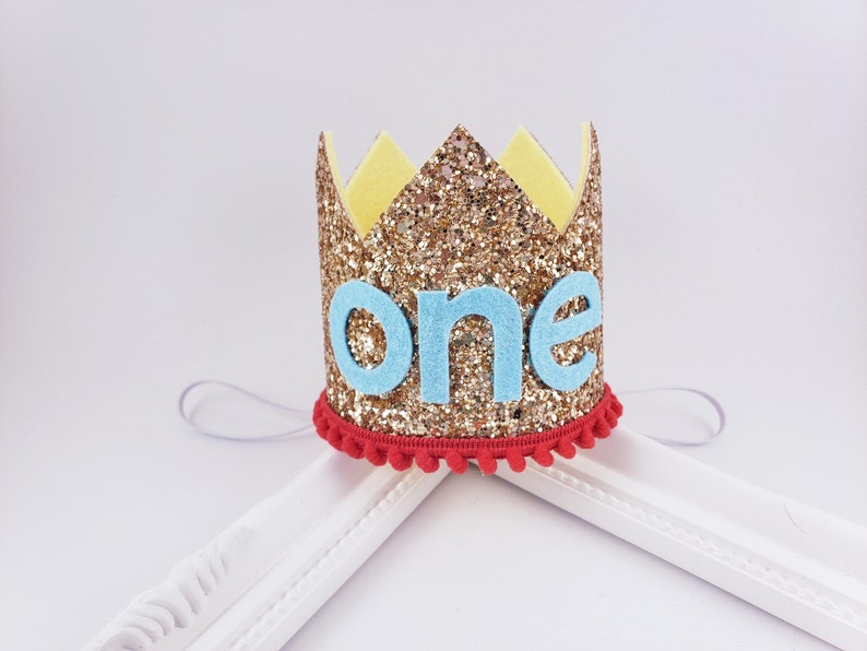 May include: Gold glitter crown with a blue felt "one" on the front and a red pom-pom trim.