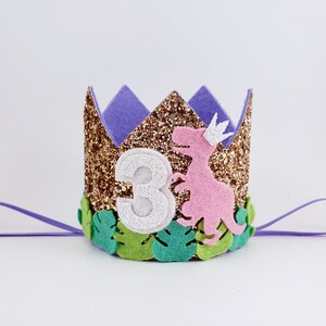 May include: Gold glitter crown with a purple felt top, a pink dinosaur, green leaves, and the number 3 in white. The crown has a purple ribbon attached.