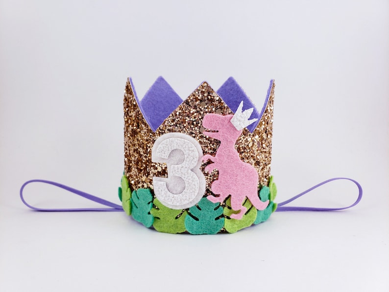 May include: Gold glitter crown with a purple felt top, a pink dinosaur, green leaves, and the number 3 in white. The crown has a purple ribbon attached.
