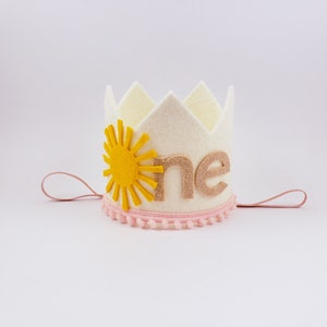 May include: A cream felt crown featuring a yellow sun design and the word "one" in tan. The crown is trimmed with pink pom-poms and has pink string ties. A celebratory accessory for a first birthday.