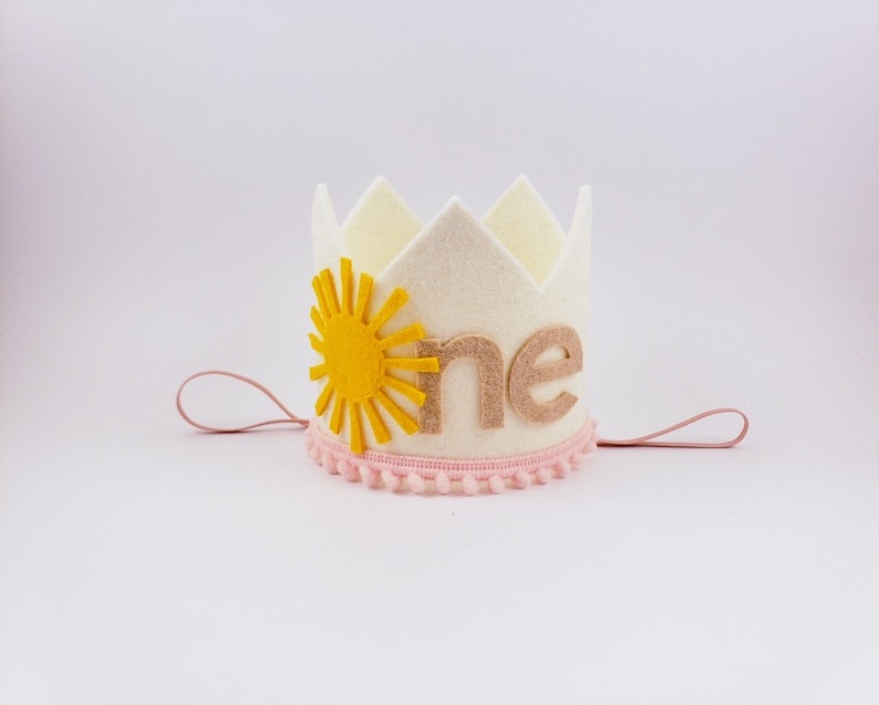 May include: A cream felt crown featuring a yellow sun design and the word "one" in tan. The crown is trimmed with pink pom-poms and has pink string ties. A celebratory accessory for a first birthday.