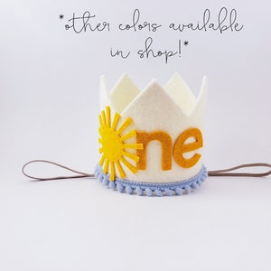 May include: A cream felt crown featuring a yellow sun design and the word "one" in gold. The crown is trimmed with light blue pom-poms and has a grey elastic band. Text in the image says "other colours available in shop!"